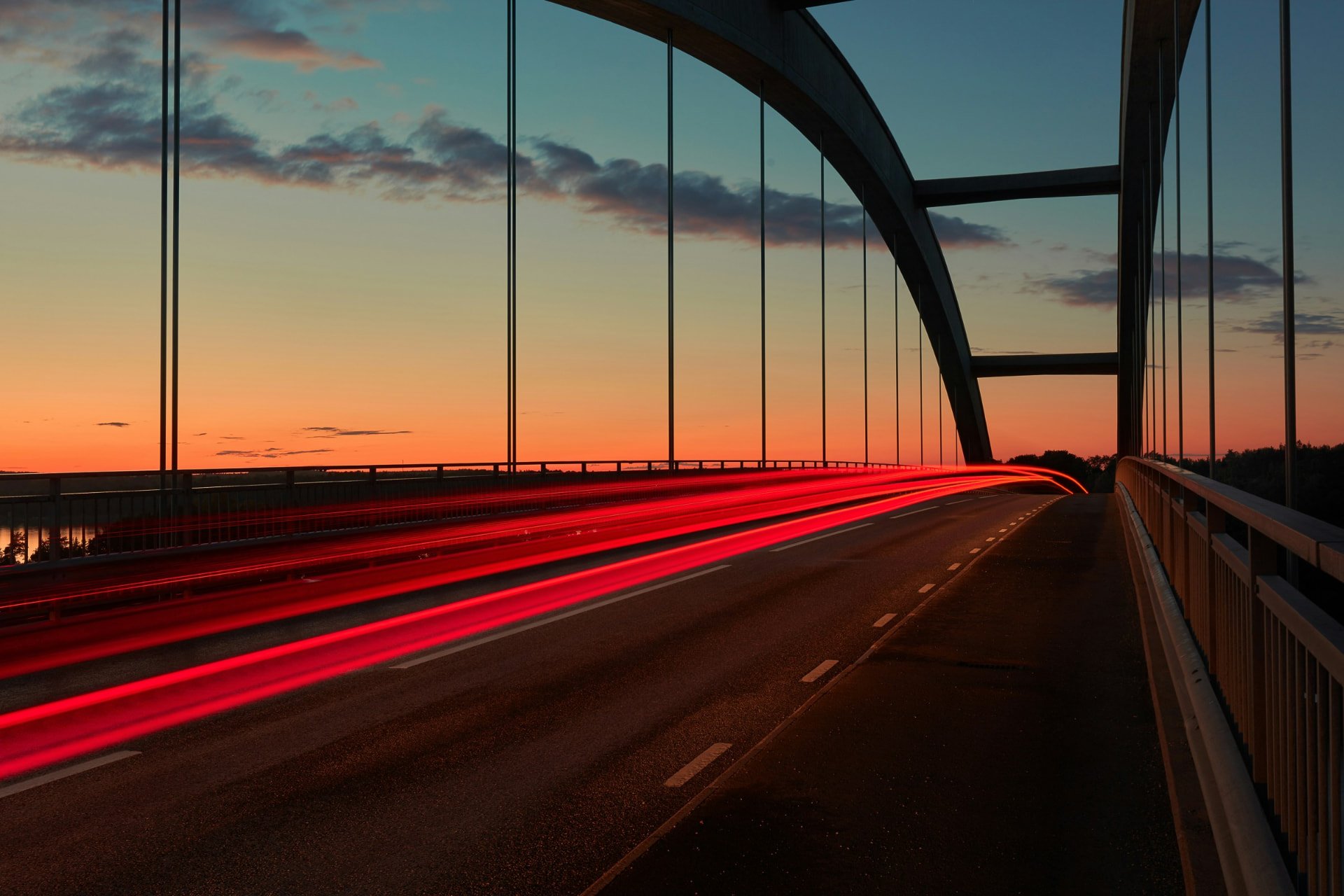 Red light trails streaming across a modern arched bridge under a sunset sky of warm orange and deep teal