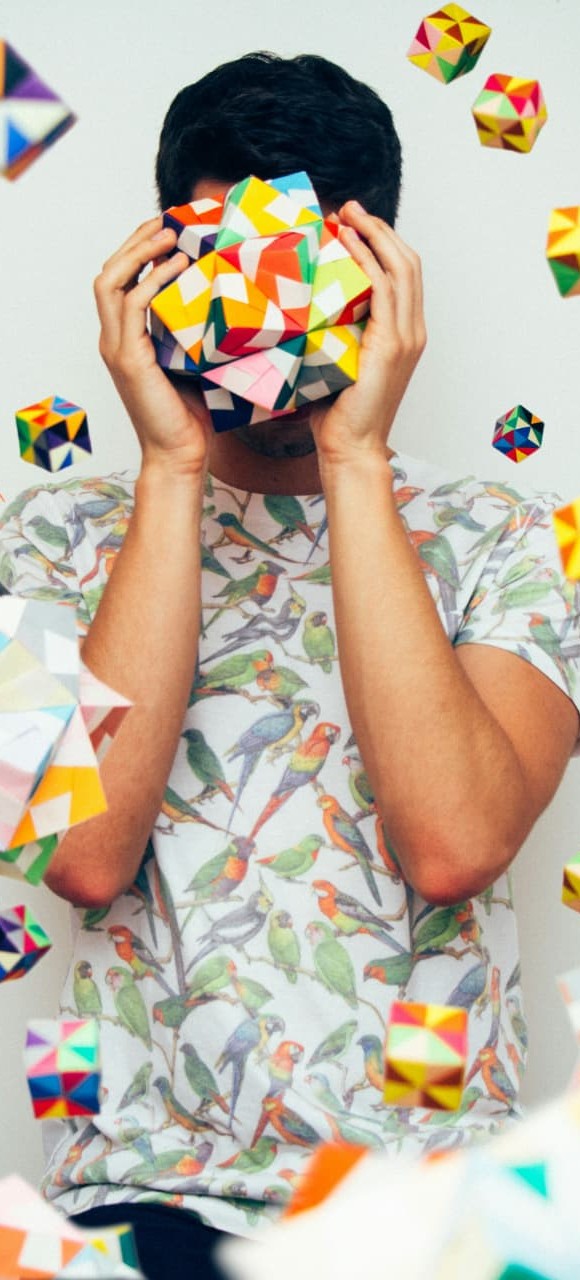 A person holding a colourful folded-paper sphere in front of their face, surrounded by floating geometric shapes
