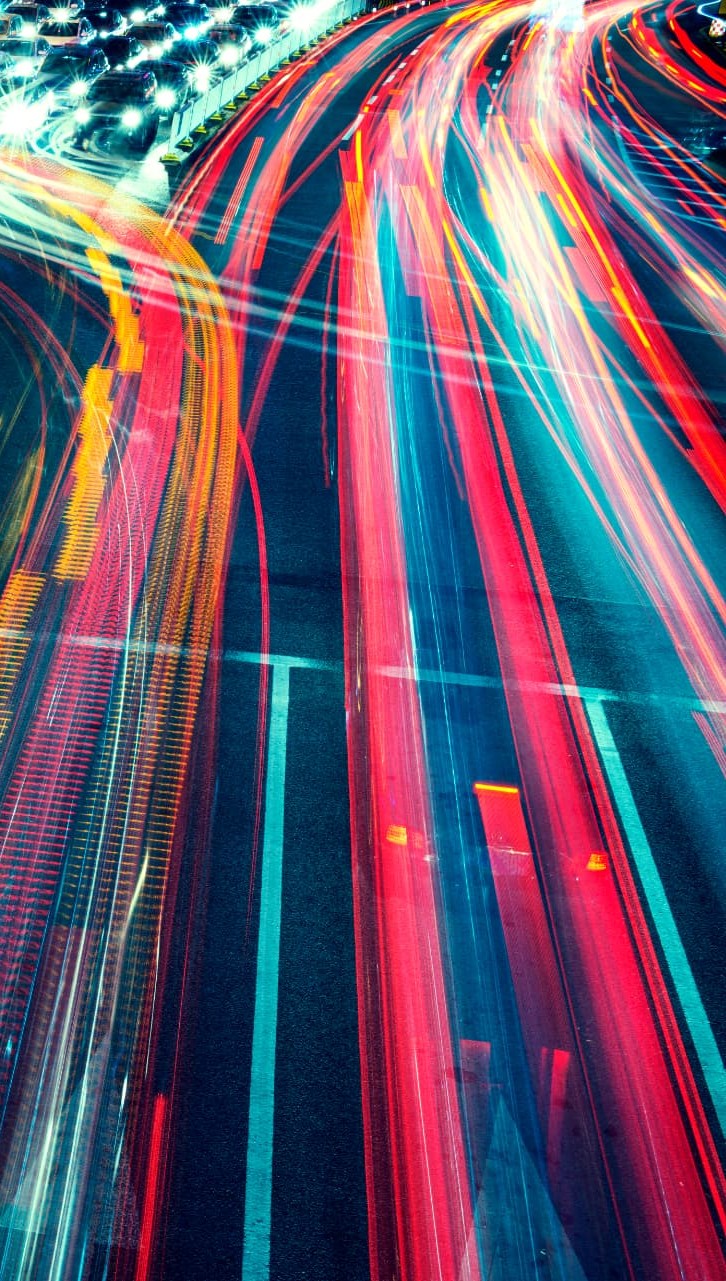 Aerial night view of a city intersection with dense vertical streams of red, cyan and golden vehicle light trails