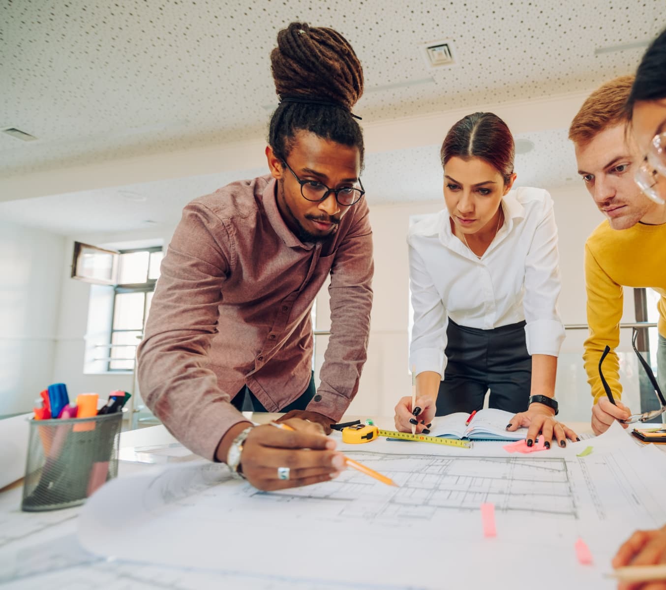 Colleagues gathered around a table of blueprints in a bright studio: a man with dreadlocks and glasses on the left, a woman in a white shirt in the centre, a man in a mustard sweater beyond them, hands and rolled plans across the table, a tall window lighting the room from the left
