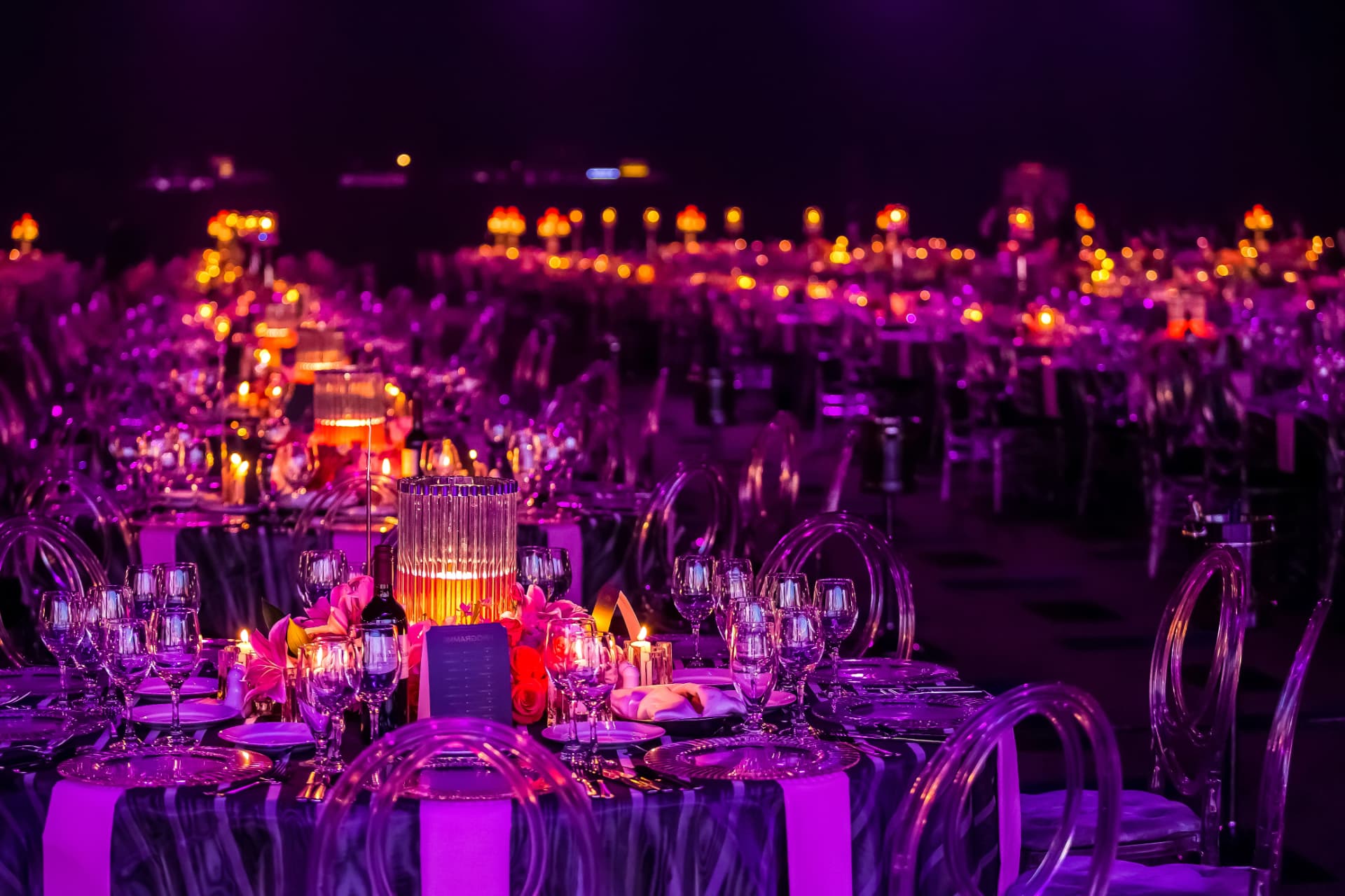An evening banquet hall: a long table dressed with wine glasses, plates and floral accents in the foreground, a tall ribbed glass hurricane lamp glowing with a candle at its centre, rows of similar tables and violet uplighting dissolving into soft bokeh behind